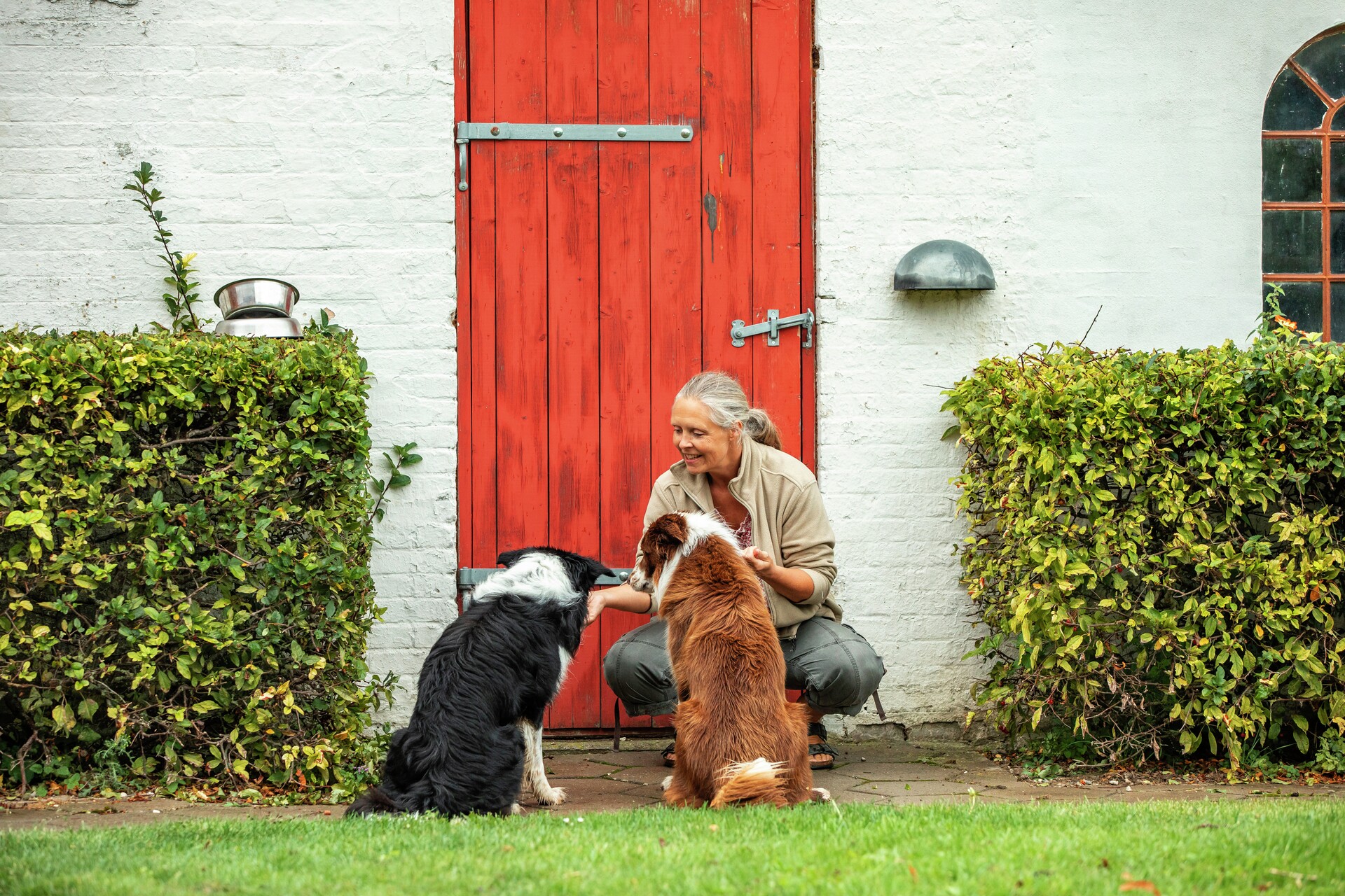Kvinde sidder på hug foran rød ladedør med to hunde, imens hun smiler, fotografi