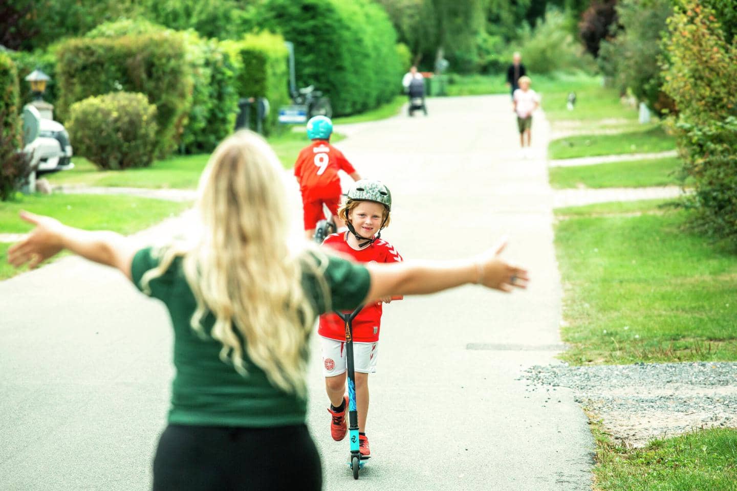 Dreng i rød trøje med cykelhjlem på løbehjul på vej mod blond kvinde med åbne arme, fotografi