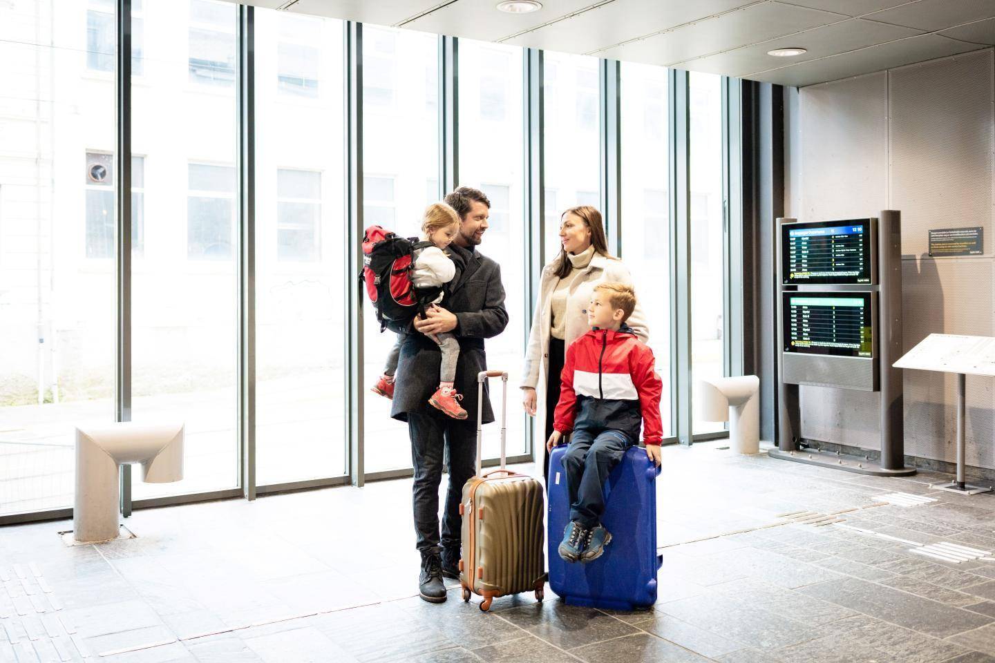 En familie står i en lys ankomsthal med kufferter foran store vinduer.